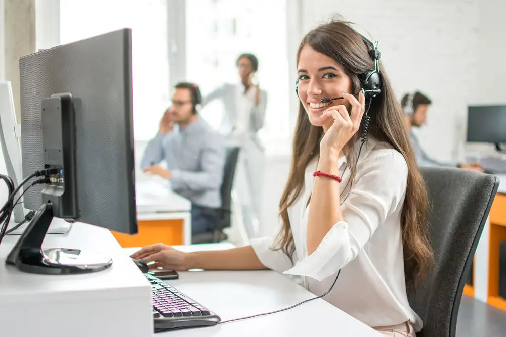 Junge Frau mit Headset am Computerarbeitsplatz in hellem Büro, Kundenservice oder Support-Situation im Hintergrund sichtbar.