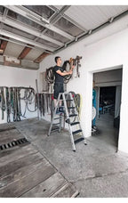 Ein mann steht auf der silbernen aluminium münk stufen-stehleiter beidseitig begehbar mit clip-step relax und hängt seile an der wand auf