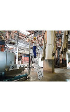 Arbeiter mit gelbem helm und blauer arbeitskleidung steht auf silberner aluminiumstufen-stehleiter munk mit clip-step r13 in industriehalle