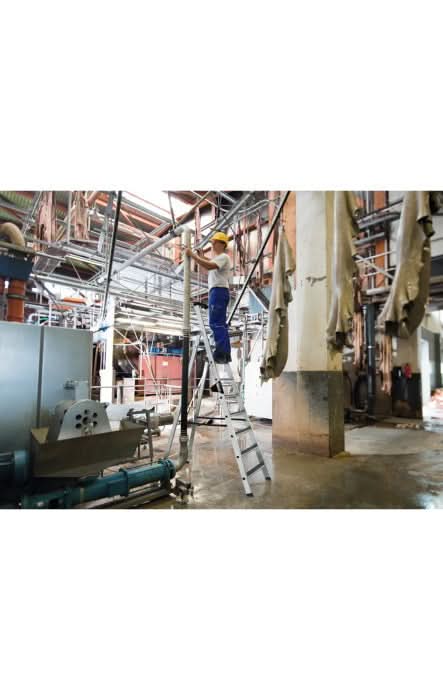 Arbeiter mit gelbem helm und blauer arbeitskleidung steht auf silberner aluminiumstufen-stehleiter munk mit clip-step r13 in industriehalle