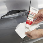 Roter und weißer sonax schleifpaste 75ml wird über einem grauen autotürgriff gehalten