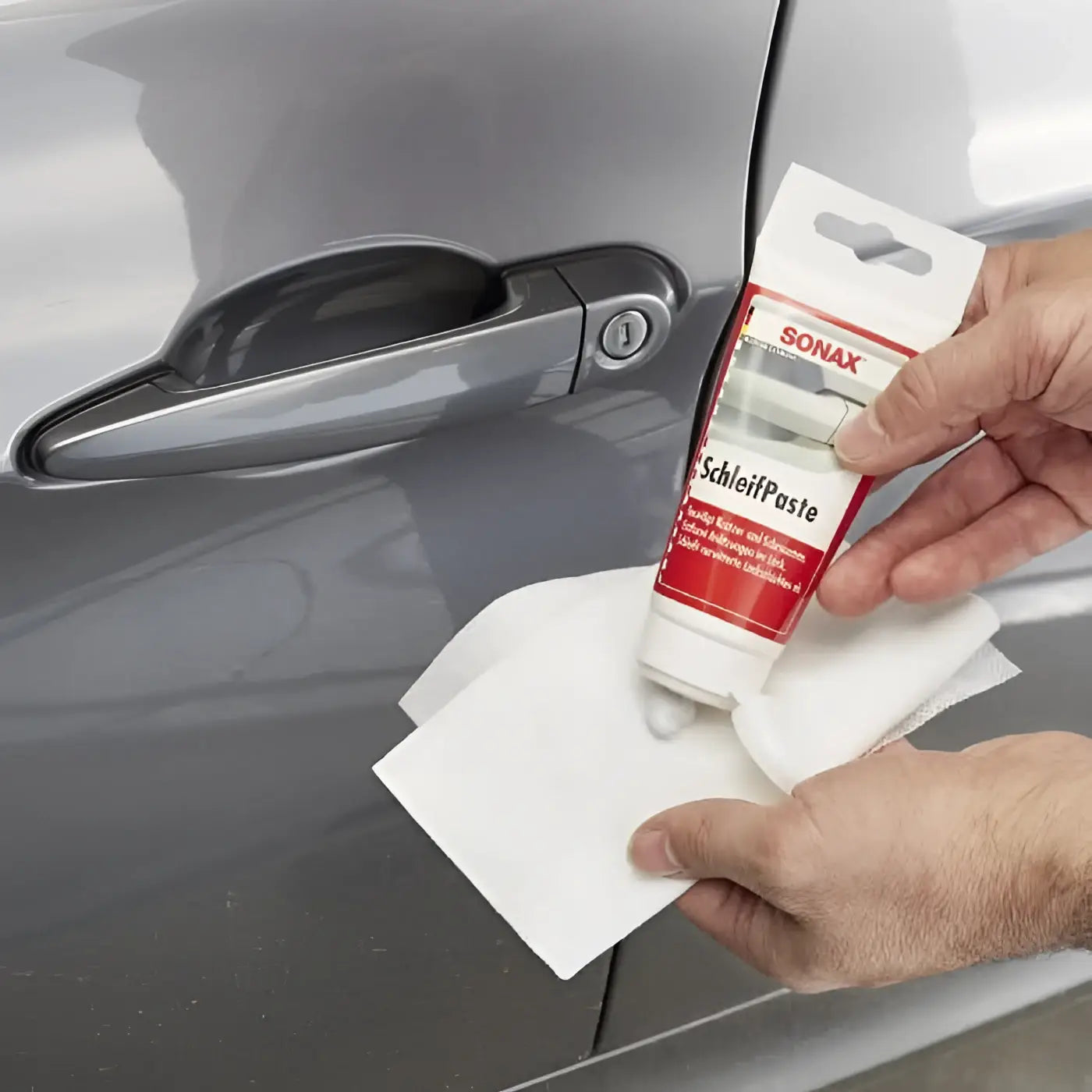 Roter und weißer sonax schleifpaste 75ml wird über einem grauen autotürgriff gehalten